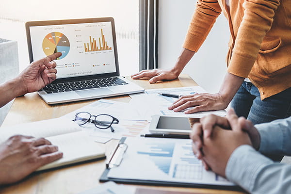 Consulenti finanziari | Immagine con persone riunite ad un tavolo pieno di appunti di lavoro mentre guardano e indicano un computer che mostra dei dati finanziari