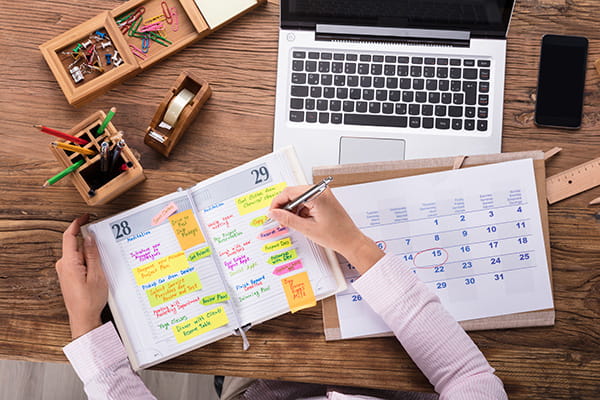 Part-time verticale | Immagine dall'alto di una persona che scrive sull'agenda, circondata da oggetti di cancelleria, da un calendario e da un computer